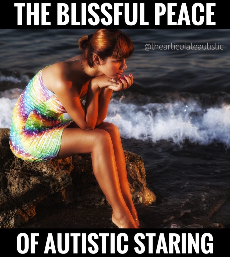 Young woman with auburn hair wearing a rainbow tank dress sitting on a rock by the ocean looking at the waves and dipping her feet into the water with text that reads, "The blissful peace of autistic staring".
