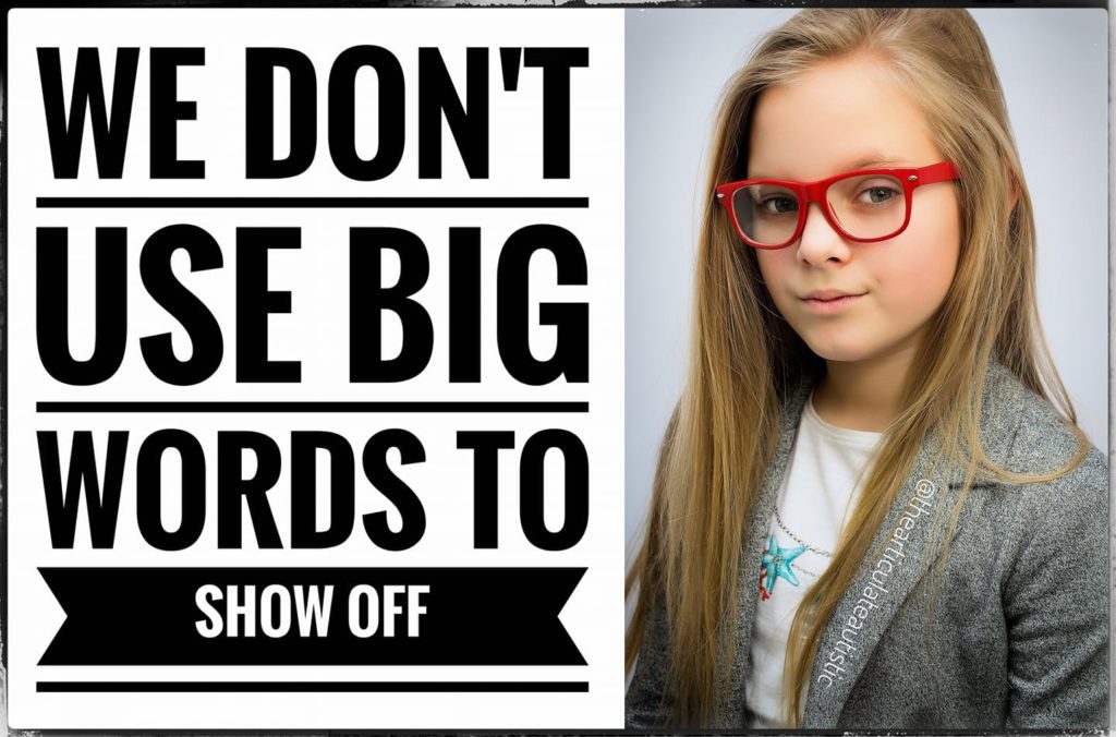 School-age girl with long, blonde hair and red glasses in a grey suit jacket with text that reads, "We don't use big words to show off".