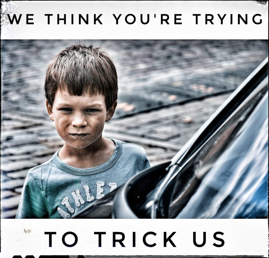 White little boy with brown hair standing outside next to a car looking skeptical (eyes narrowed, lips pursed) with text that reads, "We think you're trying to trick us".]