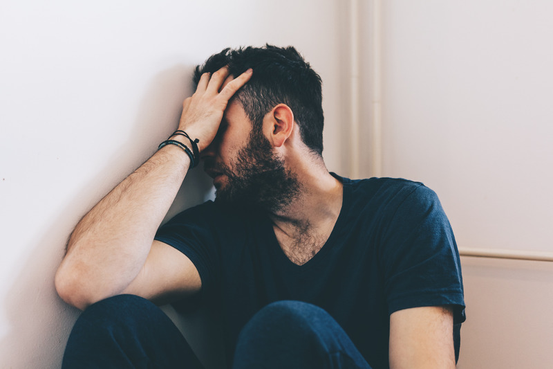 A man sitting in a corner with his face in his hands, looking upset, hurt, and/or embarrassed