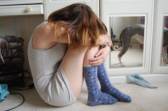 Teenage girl with blue polka-dotted socks sitting in her room with her head in her arms. She seems upset. You can see a dog reflected in the mirror next to her.