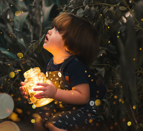 A young child holding a jar filled with LED lights. The child looks up in wonder at sparkling yellow lights surrounding him.