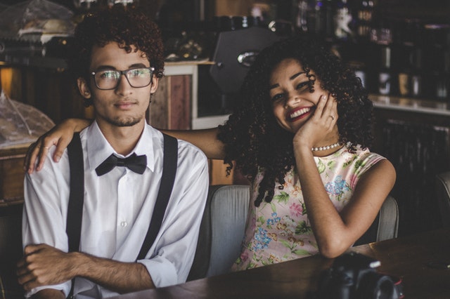 Two people sitting together in a bar. The woman, who has curly hair, is smiling with her arm around the man. The man, who is wearing glasses and a bow tie, looks at the camera with a serious look.
