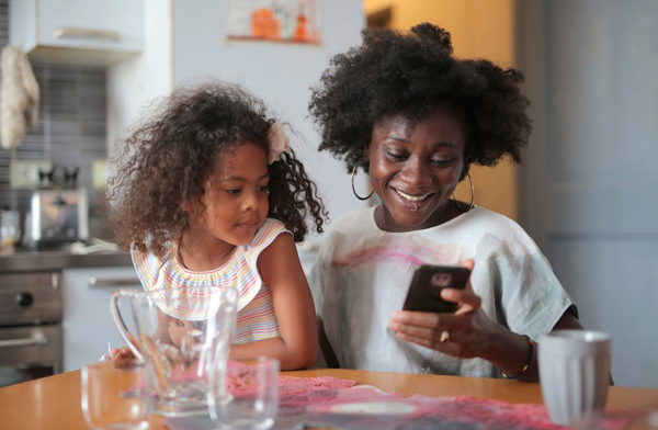 Mother and child sitting at a kitchen table, the mother is looking at her phone while her child looks on.