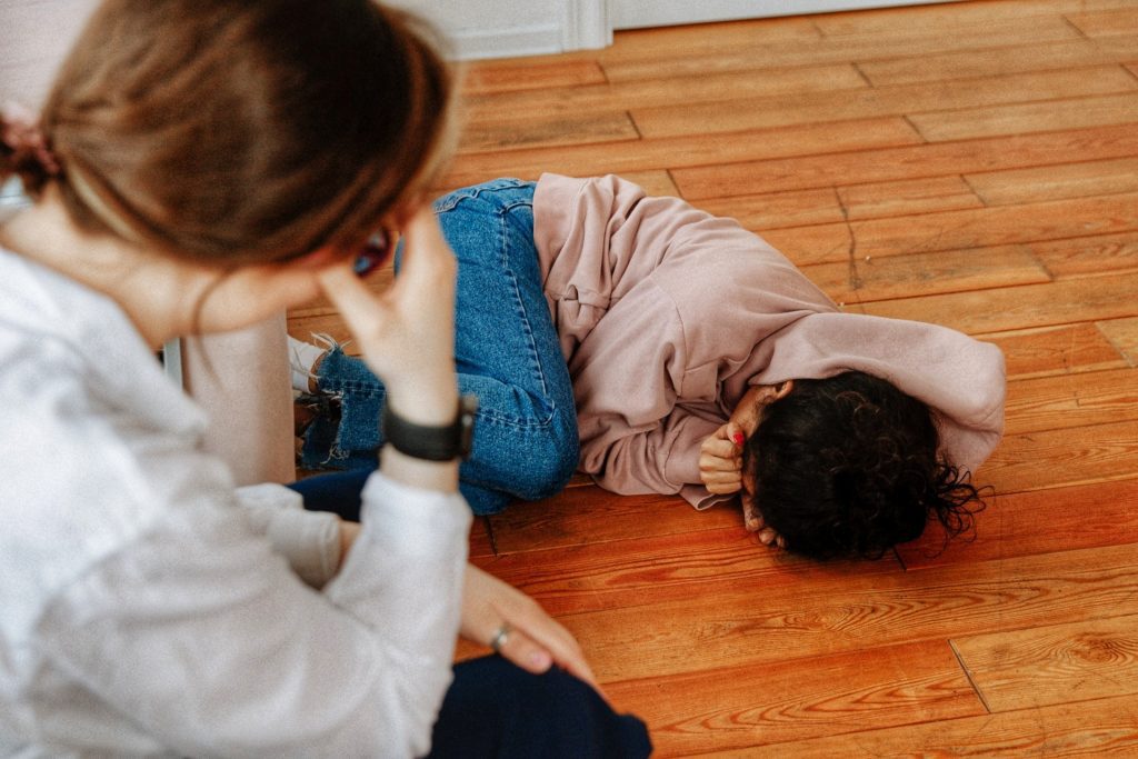 A young girl with dark hair and a hoodie curled up on the floor crying while her therapist looks on.