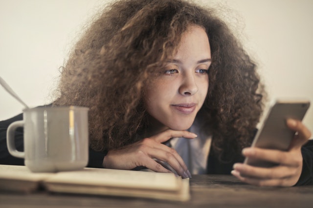 Young woman looking at her phone with a coffee cup and book in the foreground.