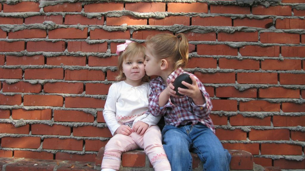 Two children sitting outside in front of a brick wall, one is kissing the other on the cheek.