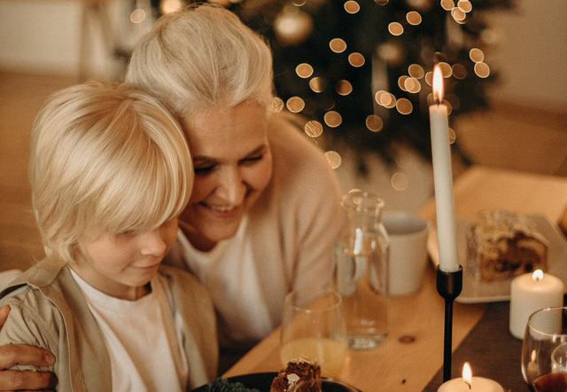 A white-haired older woman smiling and sitting close to a young blonde boy at a dinner table with holiday decorations in the background.