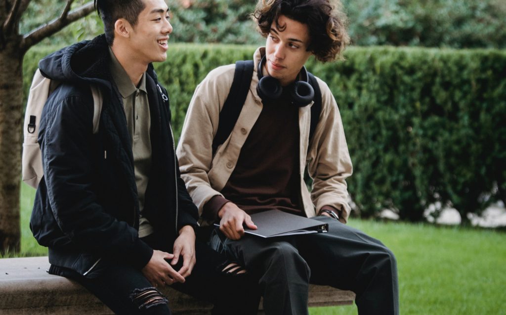 Two young men talking, one is wearing a backpack, the other has headphones resting on his shoulders.