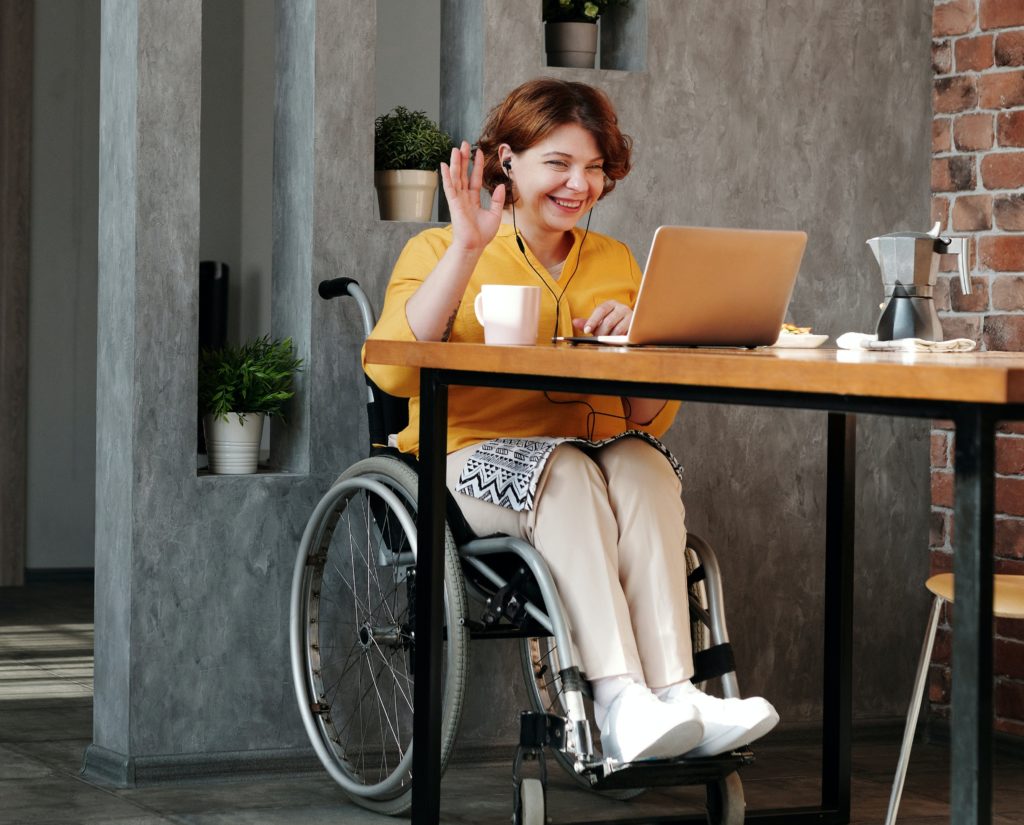 A red-headed young woman in a wheelchair sitting at her computer smiling, waving, and having a video chat. 