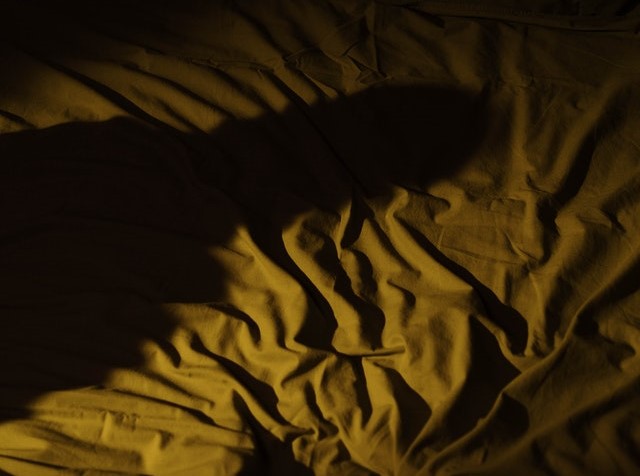 Low-light photo of rumpled bedsheets with the shadow of a person cast over the bed. 