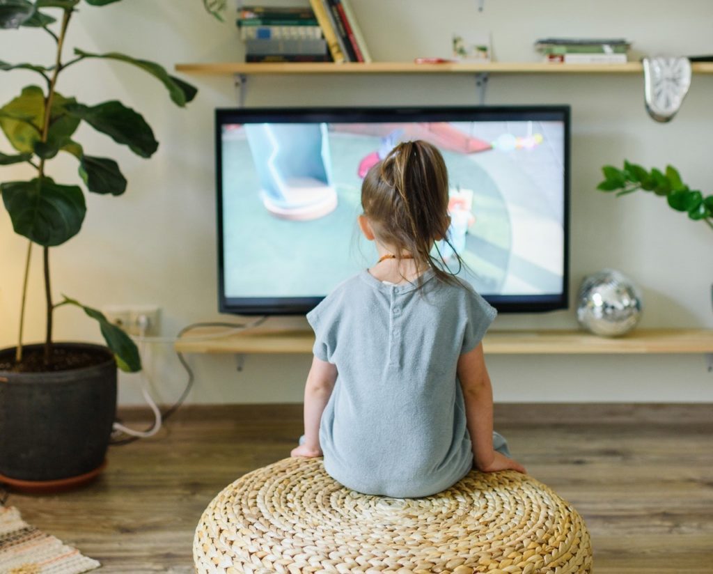 Little brunette girl watching TV with her back to the camera. 