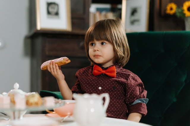 Little boy with long, brown hair, wearing a red bow tie, eating a piece of bread. 