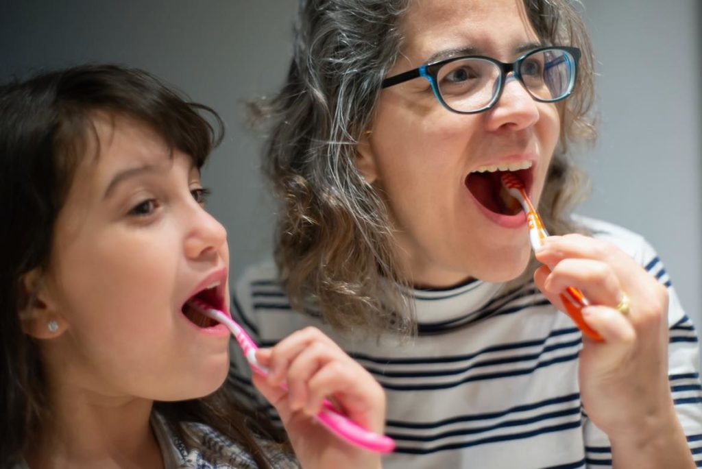 An older woman showing a young girl how to brush her teeth. The older woman is wearing blue-framed glasses, and the young girl is using a pink toothbrush. 