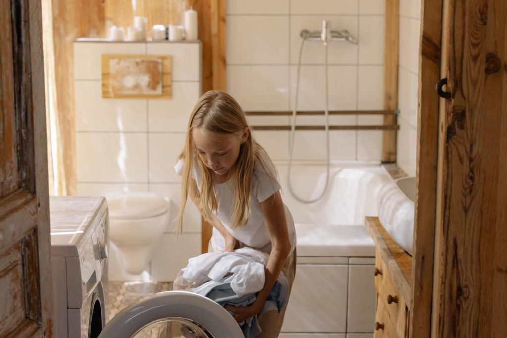 A little girl with long blonde hair doing laundry in a bathroom with a tub and toilet in the background. 