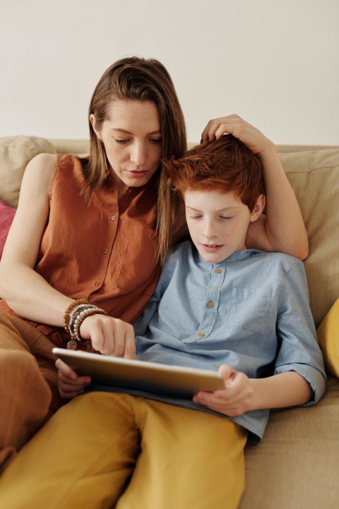 A mother and son. Son has red hair and a blue, button-up shirt. He's on a tablet, and his mother is pointing to something on the screen.