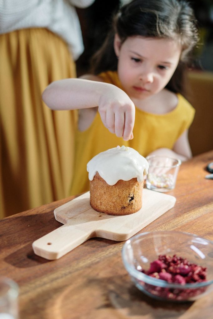 A little girl in yellow with long dark hair putting sprinkles on a cupcake while an adult looks on. 