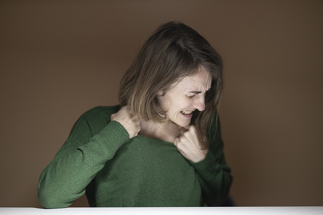 Woman with brown hair wearing a green shirt, pulling on the neck of the shirt and looking distressed and angry.