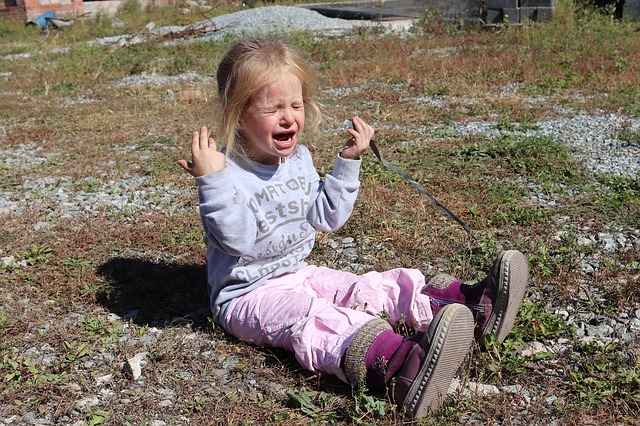 Little girl with blonde hair and pink pants sitting outside crying, her hands raised in the air.