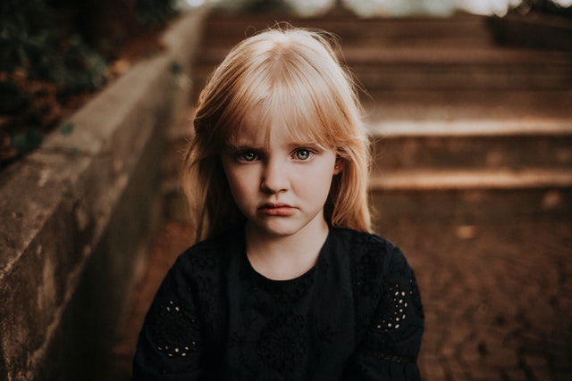 A little girl with long, blonde hair and a serious facial expression looking intently at the camera. 