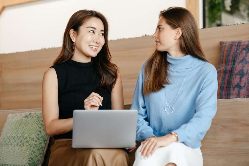 Two women having a conversation. One is holding a laptop, the other is wearing a blue sweater. Both are looking at each other. The woman with the laptop is smiling, and the woman in the blue sweater looks a bit confused.
