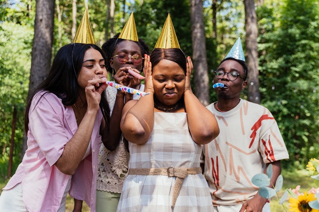 4 people wearing birthday hats. One person is covering her ears because the other 3 are blowing noisemakers near her. 