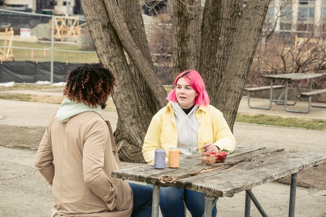 Two people sit at a picnic table together having a conversation. One has pink hair, the other has dark hair. 