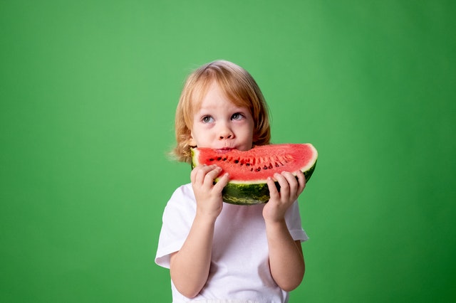 Child with blonde hair standing against a bright green background eating a large slice of watermelon. 