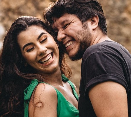 A man and woman laughing with their heads close together. The woman is wearing a green dress and turning her head to the side. Both people have their eyes closed. 
