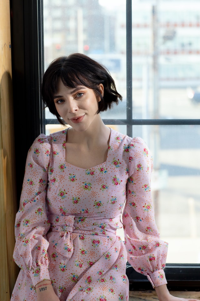 Woman with short, dark hair wearing a pink dress with red flowers. She is seated by a window.