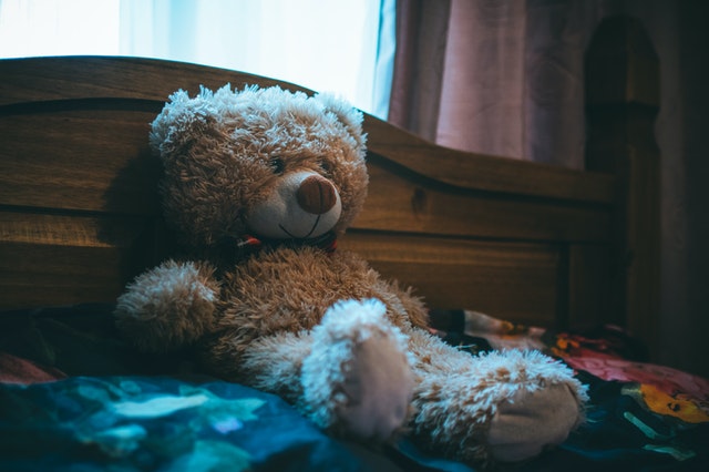 A teddy bear sitting on a bed leaning against a headboard. 