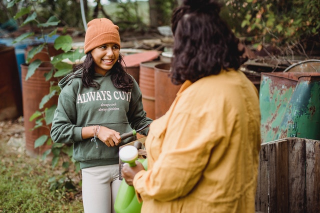 Young girl wearing a green sweatshirt and an orange hat holding gardening tools while smiling and talking to an adult woman wearing a yellow blouse. 