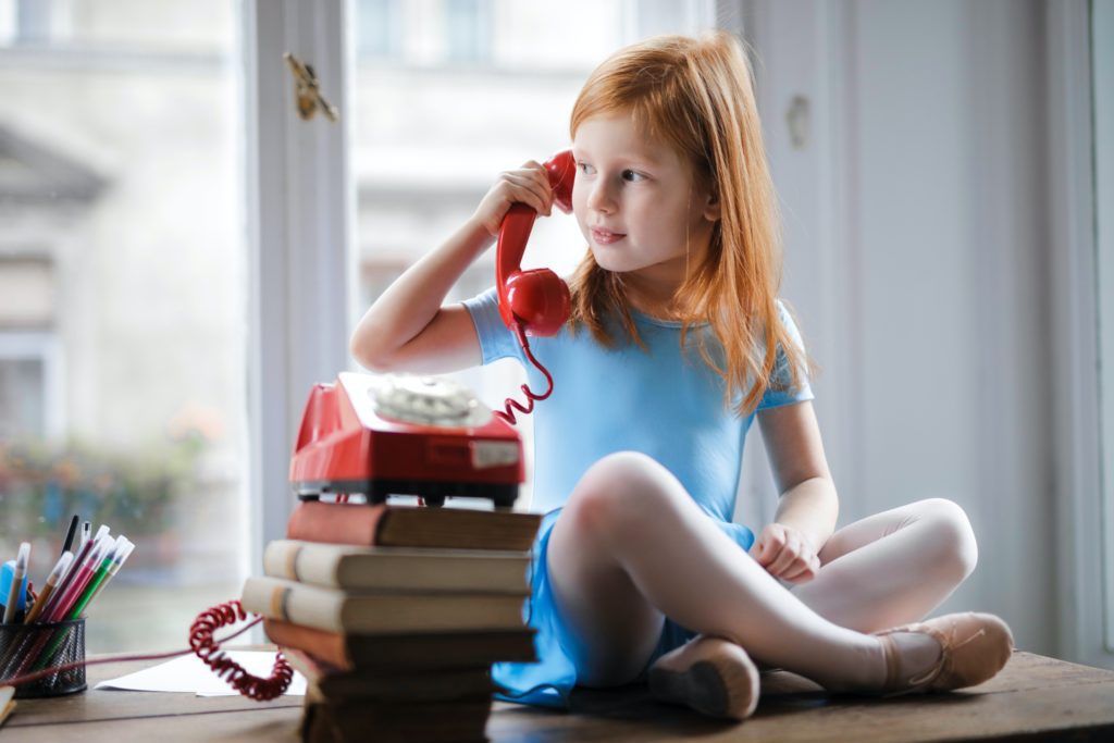 Child with red hair wearing a blue leotard "talking" on a red rotary phone while sitting on a desk. 