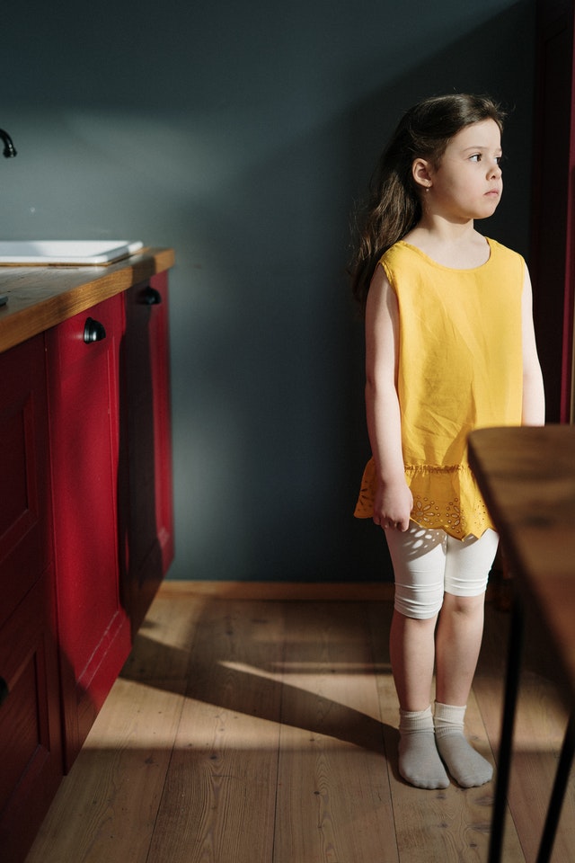 Young girl with long, dark hair in a yellow dress looks thoughtfully out of a kitchen window. 