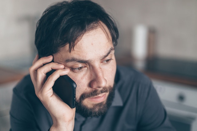 Man with dark hair and a beard on the phone, looking distressed and confused. 