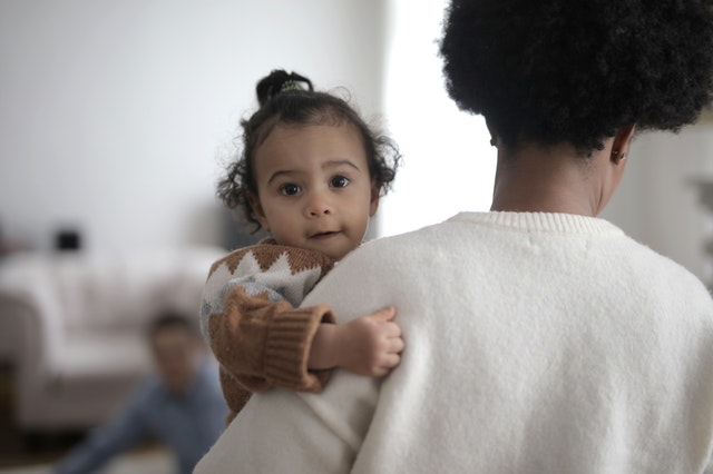 Mother holds her young child. She's wearing a white sweater, and her back is to the camera while her child looks directly at the photographer. Another child plays in the background. 
