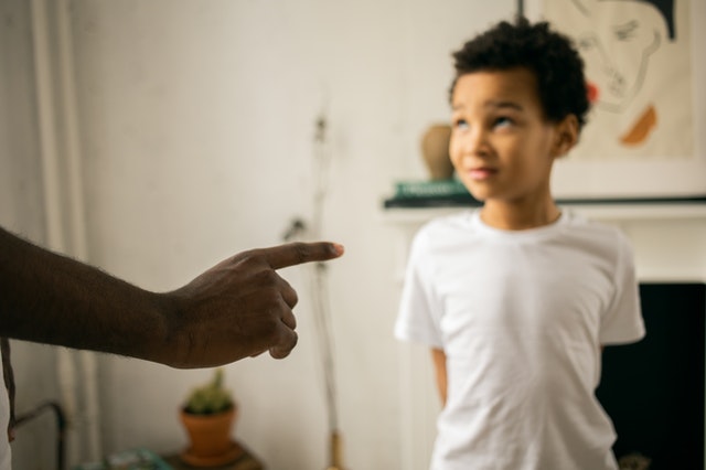 Young boy wearing a white shirt looks up at an unseen authority figure who is pointing at him with his index finger extended in a scolding manner. 