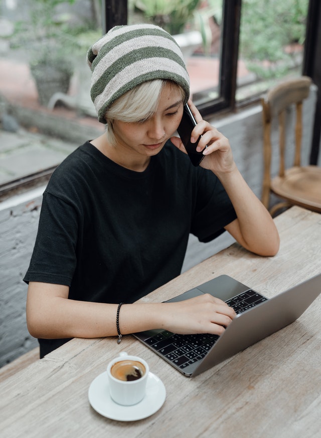 A person who appears gender non-binary sitting with a phone to their ear while looking at a laptop, a cup of coffee on the table next to them. 