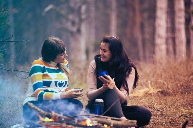 Two women talking in the woods by a fire. They've both got long, brown hair and are drinking something out of a Thermos. 