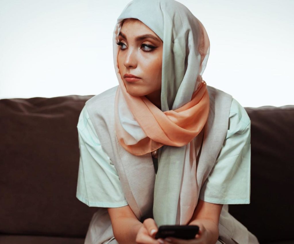 Woman with brown eyes and a colorful head scarf sitting on a brown sofa holding her phone in her hands. She looks away off into the distance. 