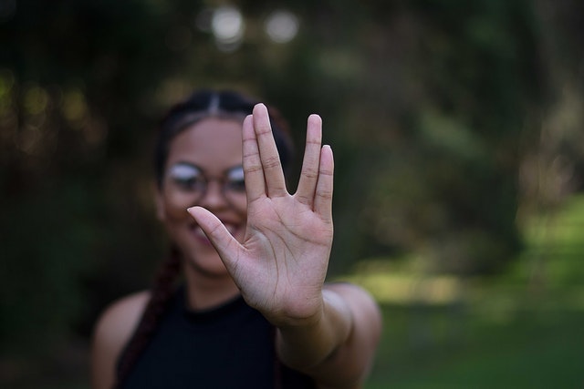 Person with dark hair and glasses holds up their hand in a classic Star Trek Vulcan hand sign with two sets of fingers together on either side, forming a 'V'. She is smiling. 