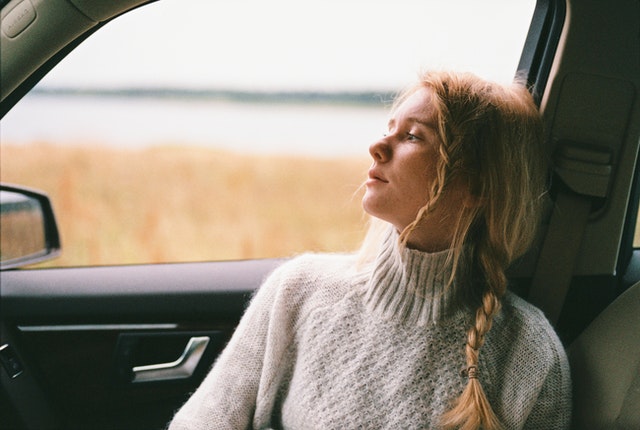 Young blonde woman in a white sweater sitting in a car, looking out the window with a contemplative expression. 