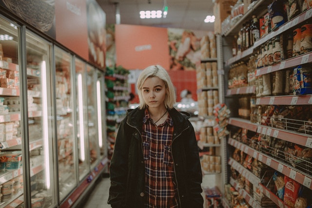 Teenager is oversized clothes standing in an grocery store aisle, her facial expression serious. 