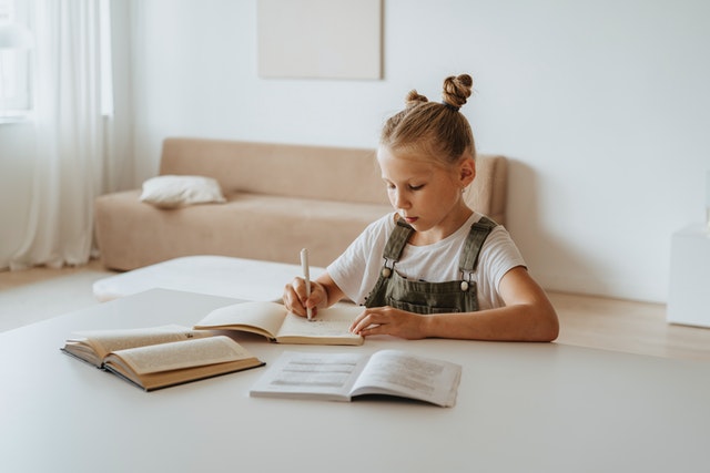 Little blonde girl with her hair up in pigtails doing homework at a desk or table. 