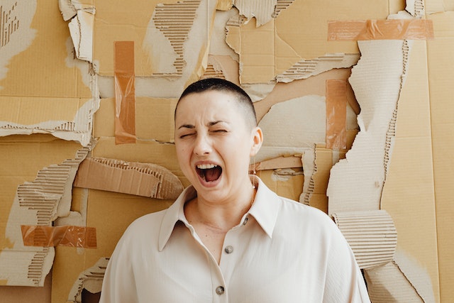 Woman with very short hair and an off-white button-down shirt standing next to a cardboard wall and screaming.