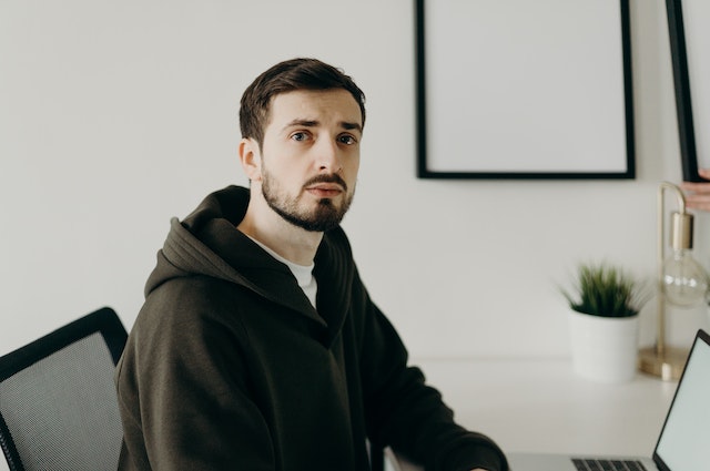 Young man with dark hair and a dark sweatshirt looking directly at the camera. He's bearded and wears a neutral or confused facial expression. 