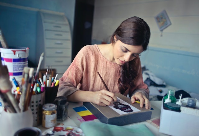 Woman with long, auburn hair in a ponytail tucked over her left shoulder. She's wearing a light-weight, peach-colored shirt and painting/drawing. 