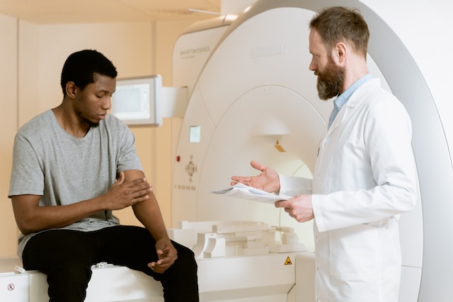 A patient sitting on an MRI/CAT scan machine holding his left arm with his right while a doctor or technician stands in front of him with papers in his hand. 
