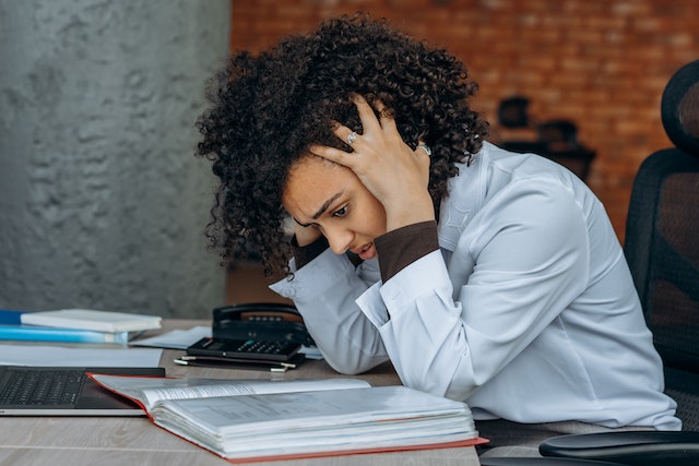 Woman with curly hair and a white blouse holding her head in her hands in frustration while she looks at an open book.