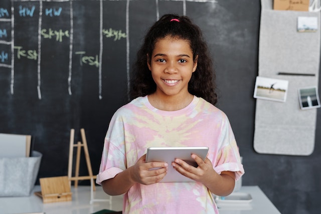 Happy, smiling little girl wearing a tie-dyed shirt while standing in front of a blackboard and holding a tablet. 